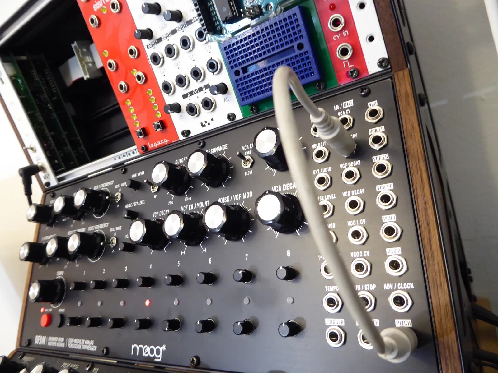 A Moog DFAM mounted below a Eurorack case, patched with a grey cable. Knobs for two oscillators, noise, filter cutoff and resonance and the decay envelopes sit above two rows of eight step knobs, with a patch bay of jacks on the right.
