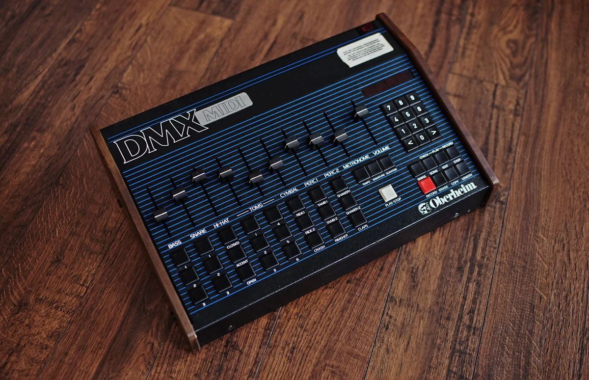 An Oberheim DMX on a wooden table: a black and blue panel with a row of voice faders, a numeric keypad and a red record button.