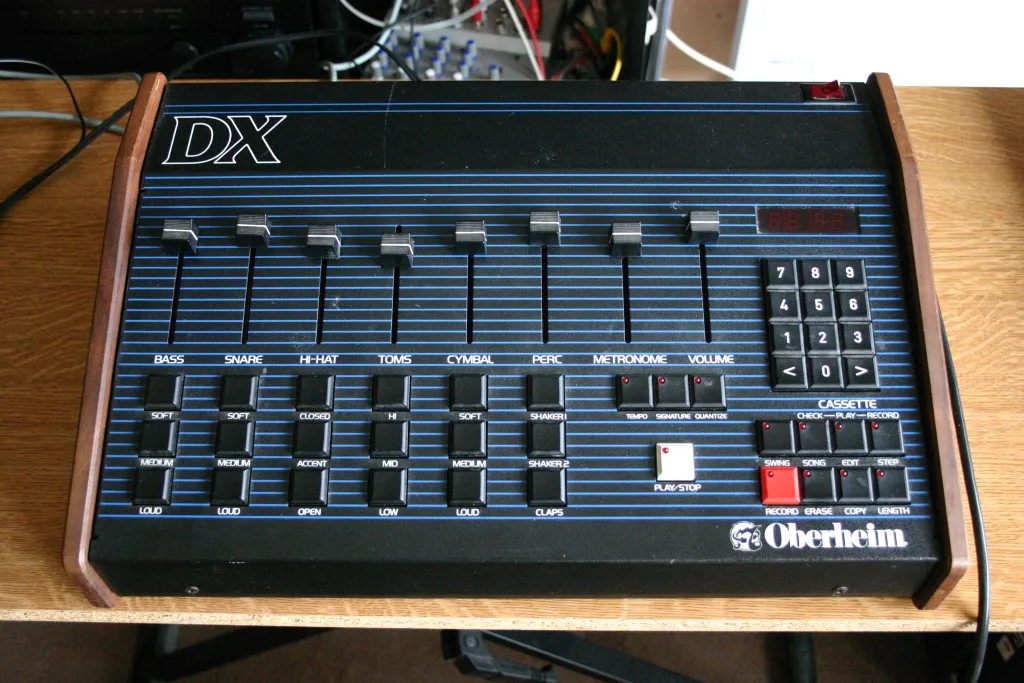 Oberheim DX front panel with six voice faders for bass, snare, hi-hat, toms, cymbal and percussion plus metronome and volume, a four-digit red LED display and lighted function buttons, framed in imitation wood end cheeks.