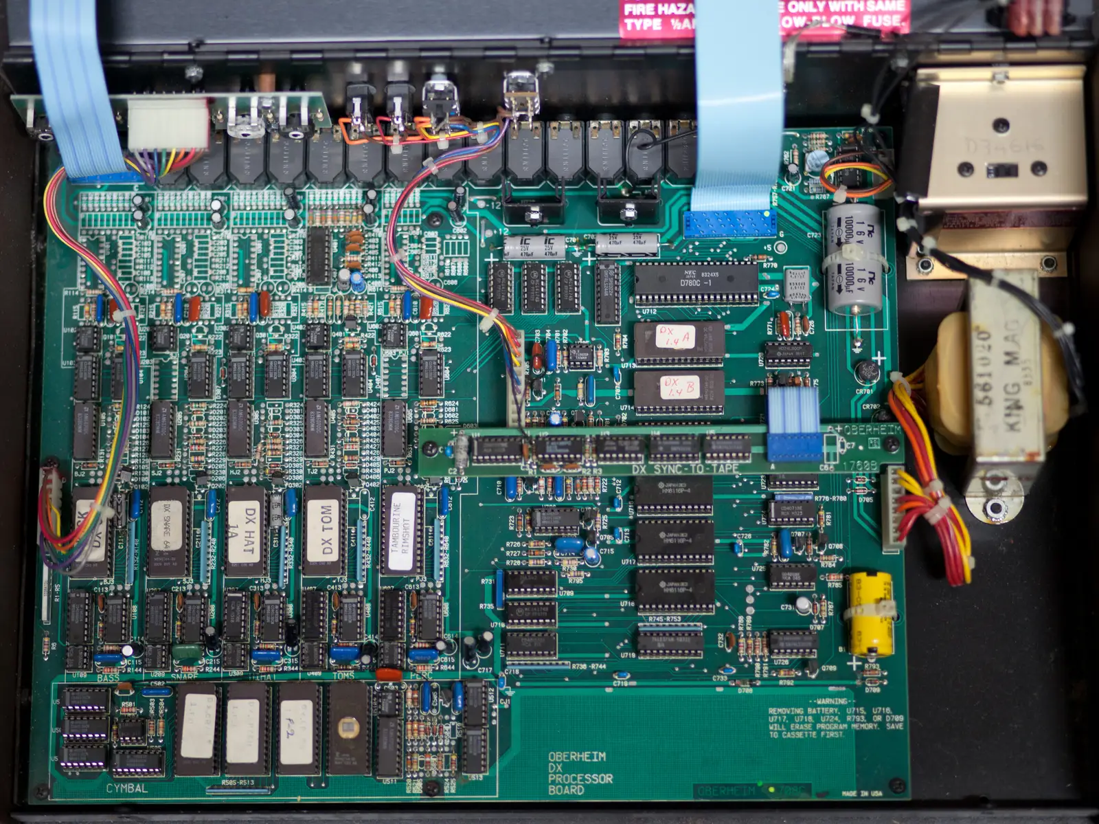 Open Oberheim DX showing its green processor board. A row of socketed EPROM chips with handwritten labels such as DX HAT and a tambourine and rimshot chip sits above the bass, snare, toms and cymbal sections, with the output jacks along the top edge.