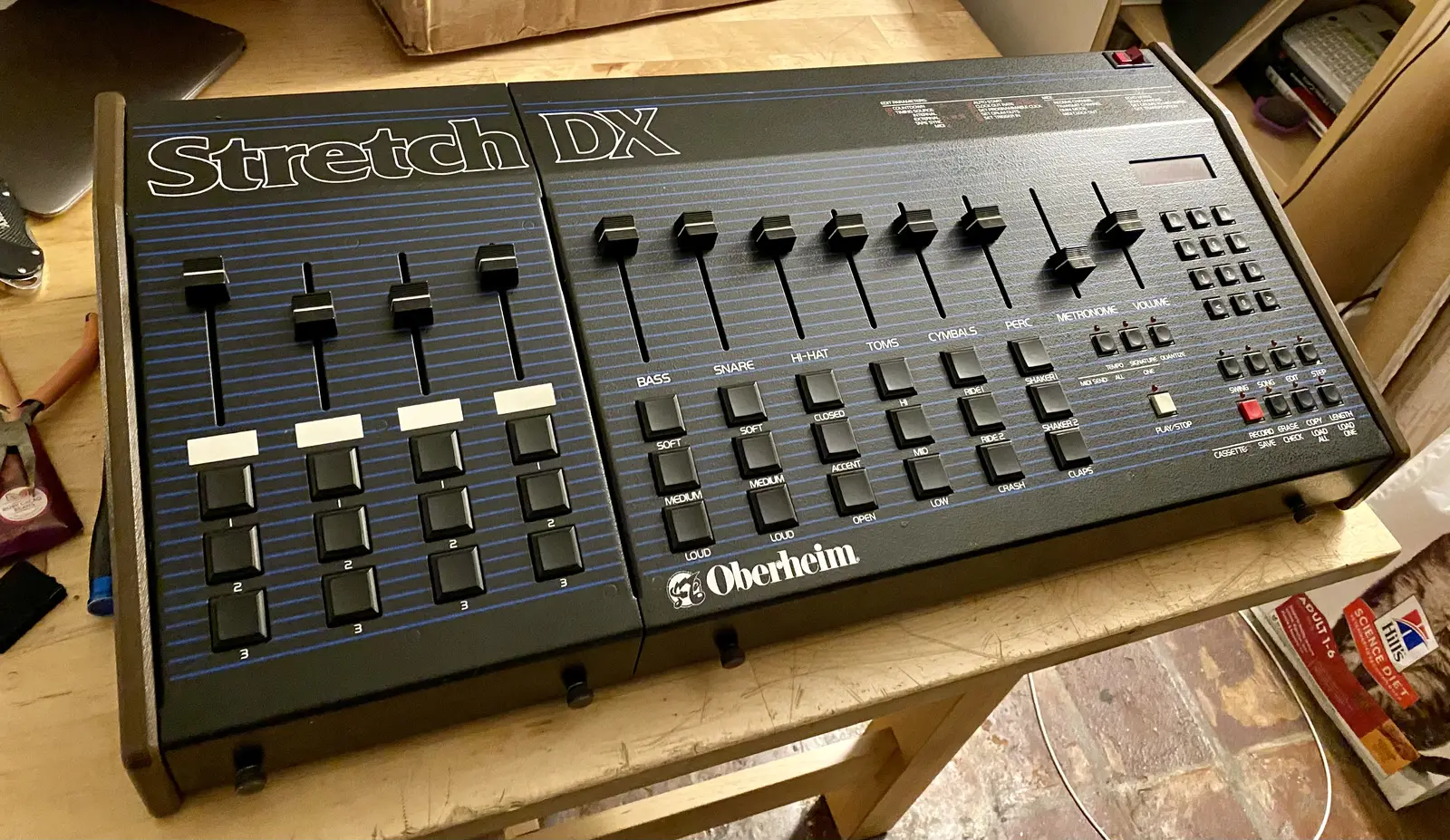An Oberheim Stretch DX: a standard DX on the right with a matching add-on panel bolted to its left side, carrying four extra faders, each above a column of three sound buttons with blank white labels.