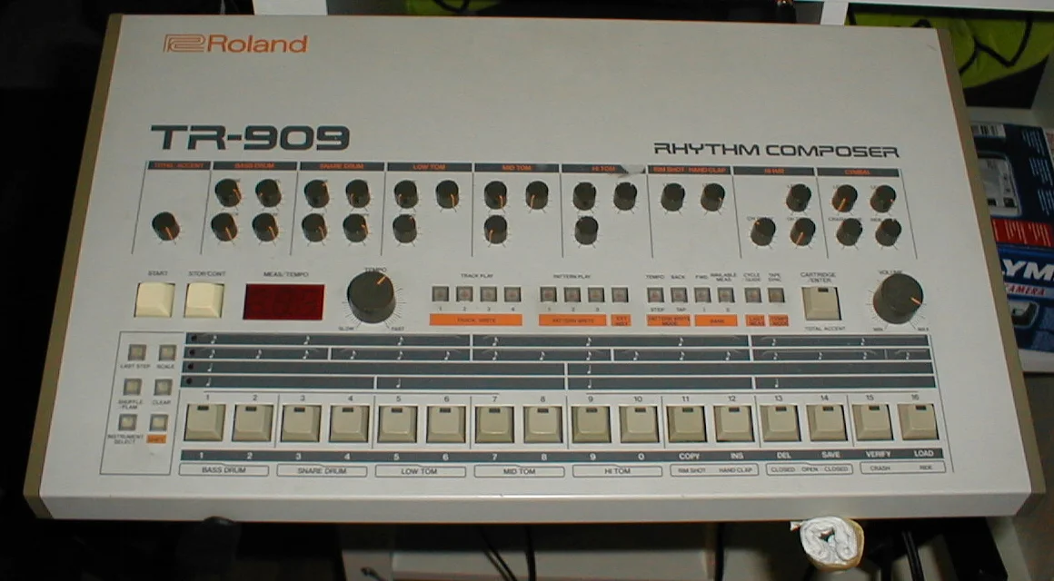 Top panel of a TR-909 on a studio stand, showing the labelled voice sections left to right: Total Accent, Bass Drum, Snare Drum, the three toms, Rim Shot and Hand Clap, Hi Hat and Cymbal.