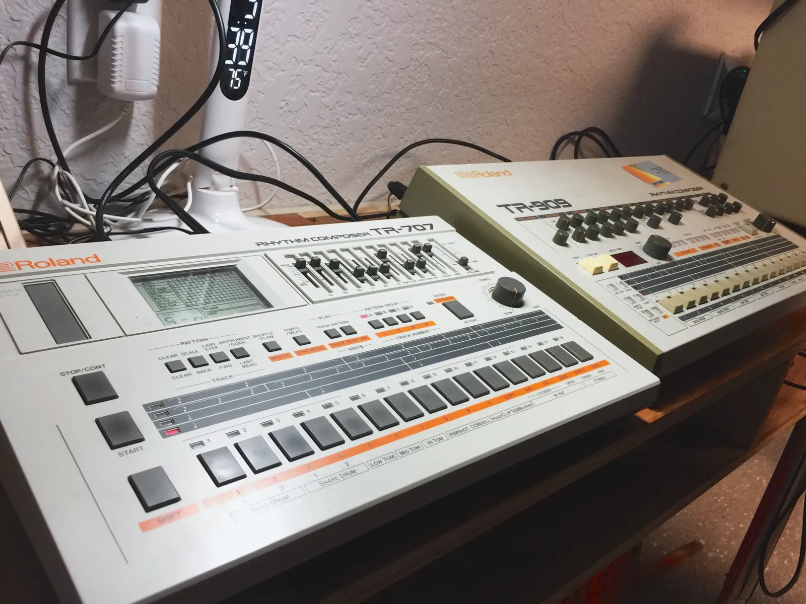 A Roland TR-707 in the foreground with its slider mixer and cartridge slot, and an original TR-909 behind it on the same desk, cables running to both.