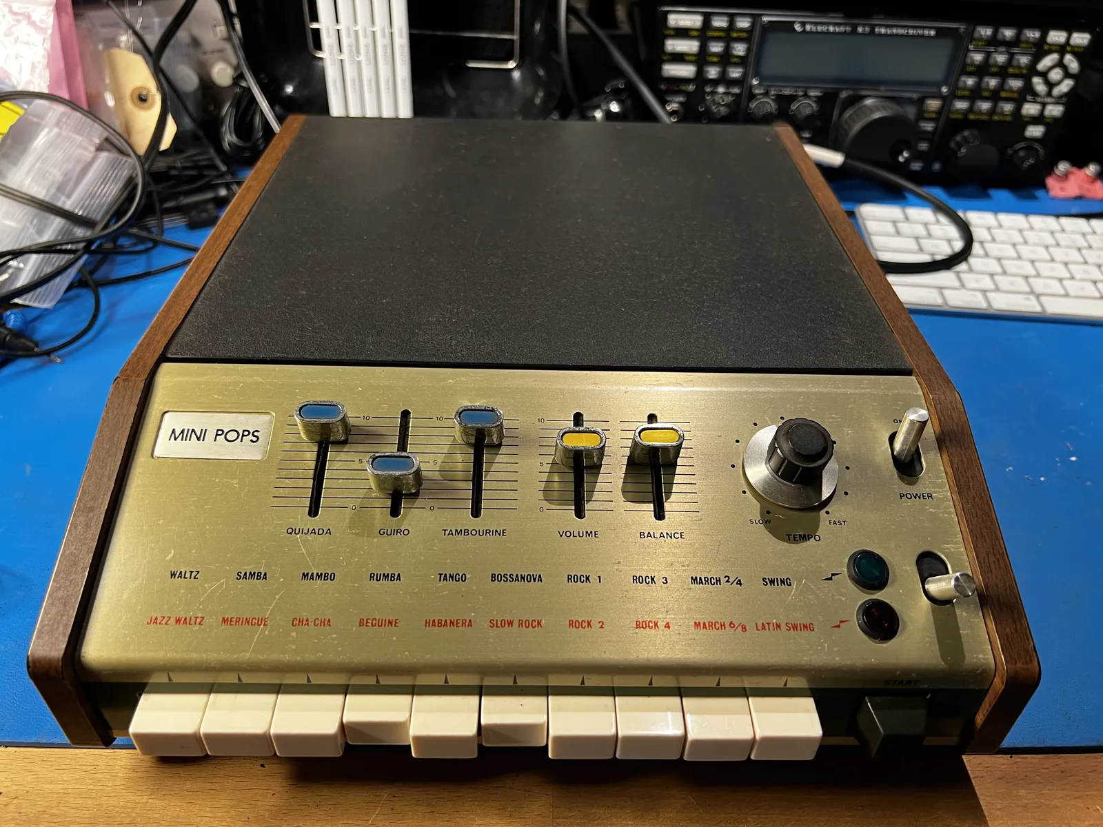 A Korg Mini Pops 7 in a wooden case: sliders for quijada, guiro, tambourine, volume and balance, a tempo knob, and twenty rhythm names from waltz to latin swing above a row of white selector keys.
